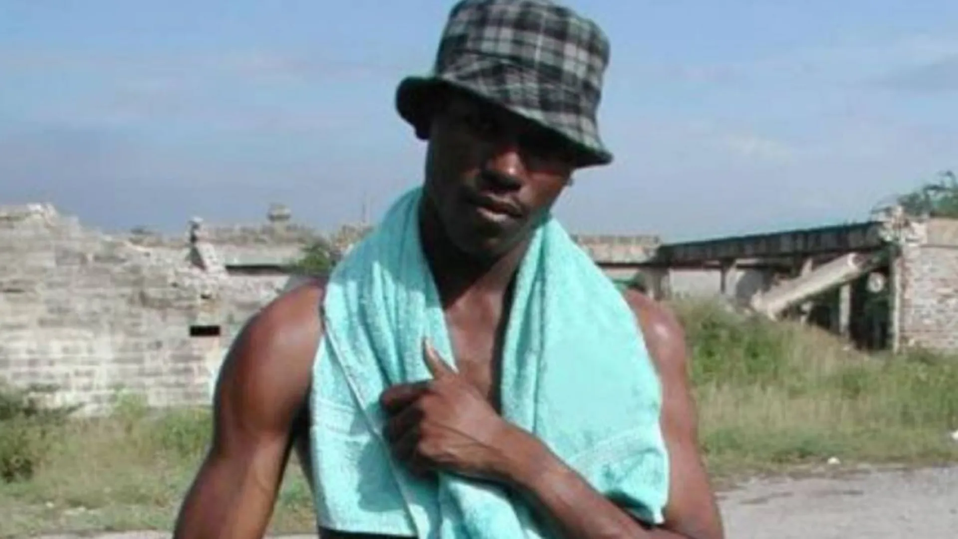 Man wearing a plaid bucket hat and holding a light blue towel over his shoulder, standing outdoors in front of old stone ruins and grassy area under a clear sky